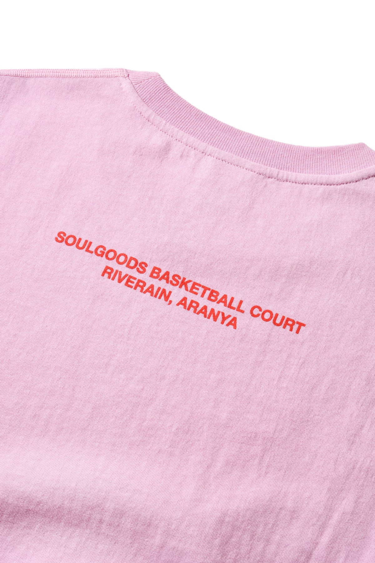 Close-up of a light pink crewneck t-shirt with red embroidered text reading 'SOULGOODS BASKETBALL COURT RIVERAIN, ARANYA' on the upper chest.