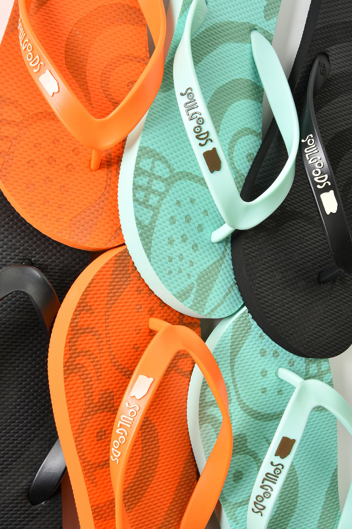 A close-up flat lay of multiple Havaianas flip-flops in orange, mint green, and black, arranged diagonally. The straps feature the white soulgoods logo