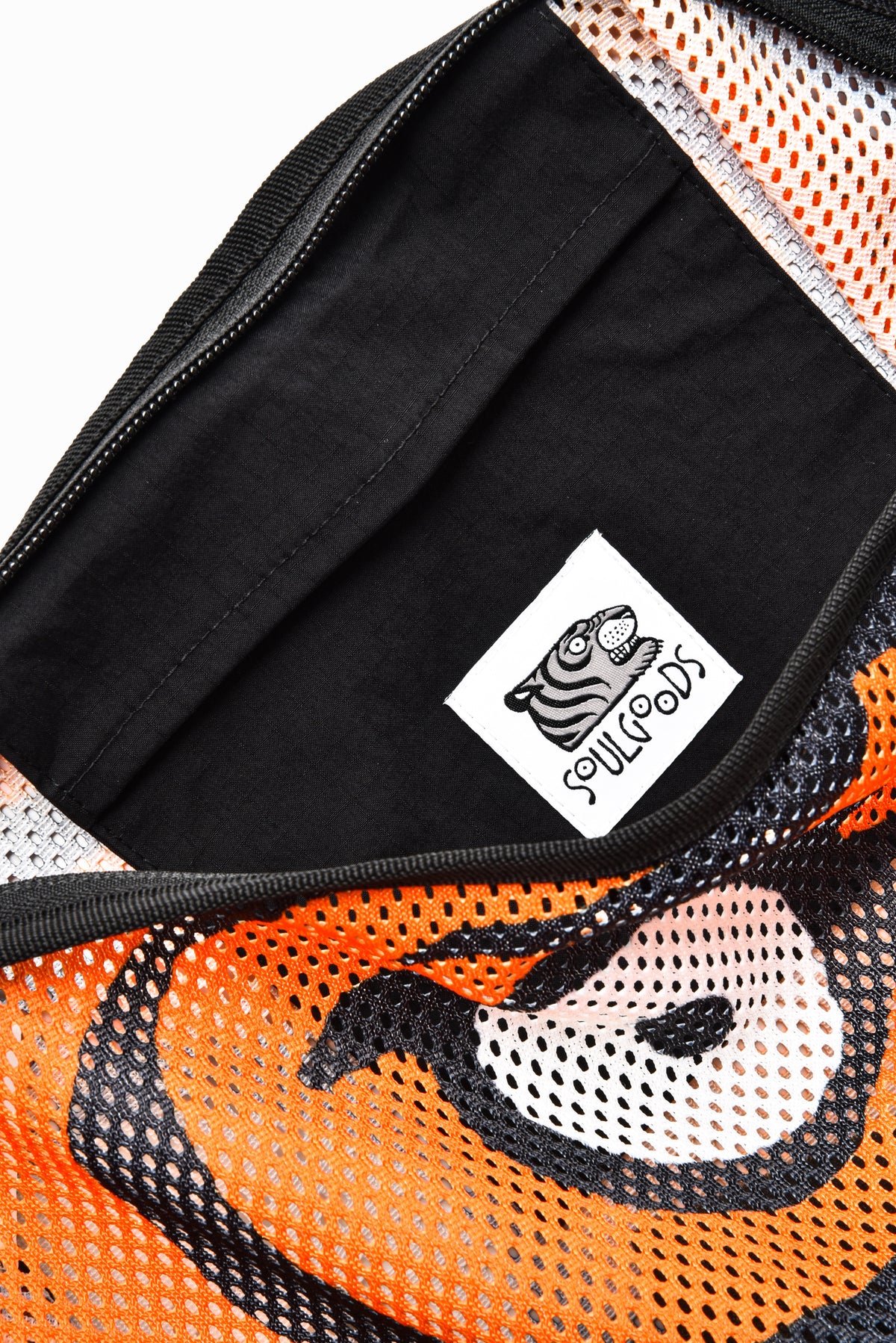 Close-up of a black zippered pouch or bag interior with a white sewn-on label reading 'SOULGOODS' and a stylized tiger face graphic. The exterior visible at the edges features orange mesh fabric with black and white tiger-face patterning.