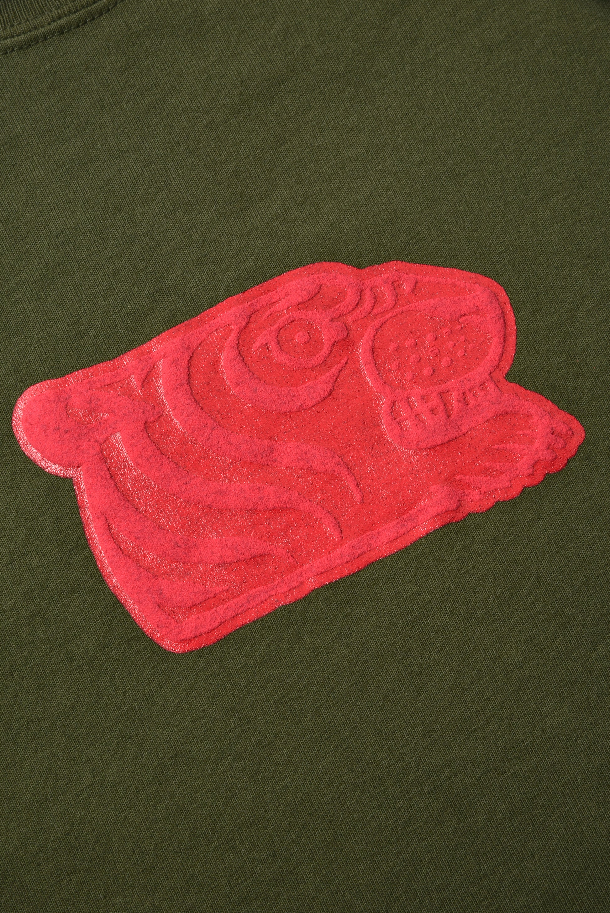 A close-up of a bright pink embroidered patch with a stylized cartoon face and swirl details, applied to an olive green fabric surface.
