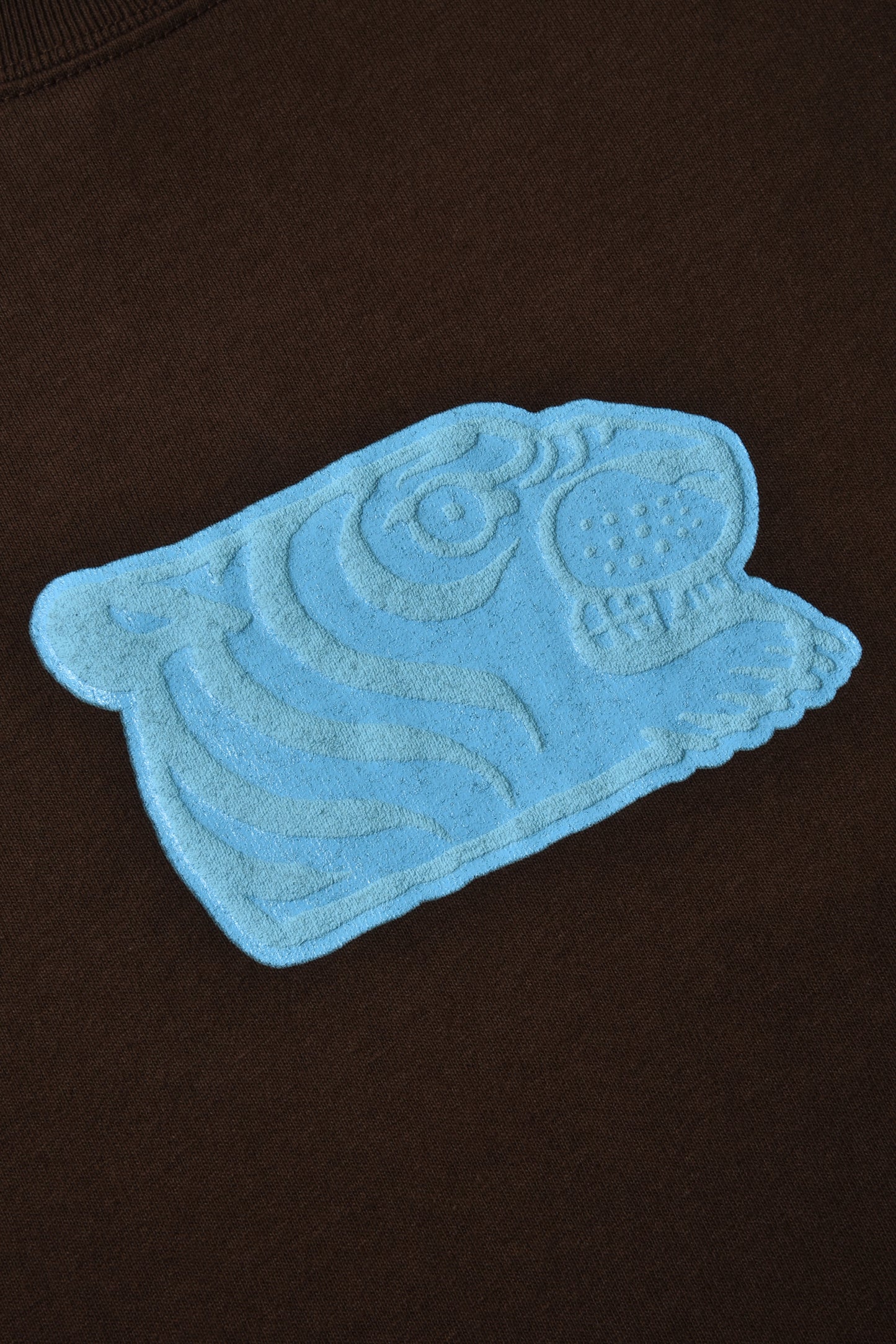 A light blue embroidered patch shaped like a stylized animal head with a circular eye and textured fur details, applied to a dark brown fabric background.
