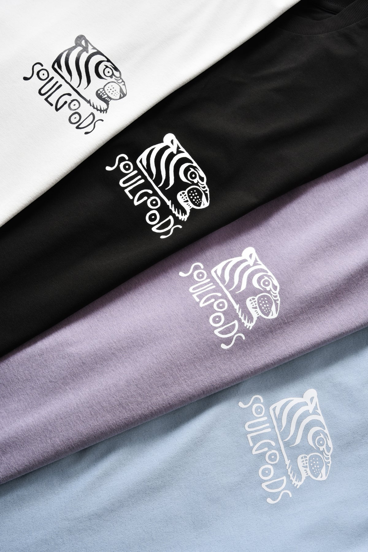 Four folded T-shirts in white, black, lavender, and light blue, each printed with a white tiger head logo and the word 'SOULGOODS' in a stylized script.