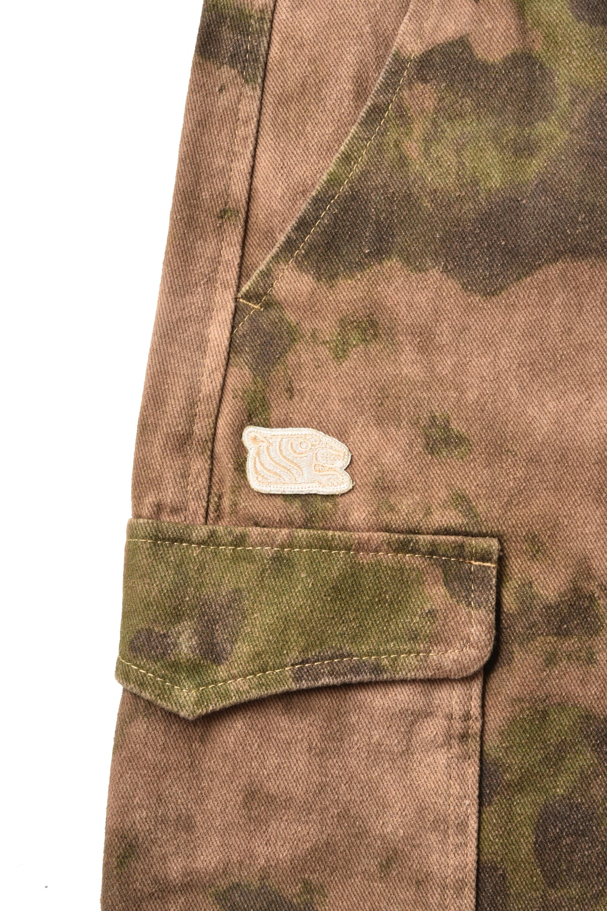 Close-up of a camouflage jacket or overshirt showing a brown and green camo pattern, a flap pocket, and a small beige tiger head embroidered patch on the chest.