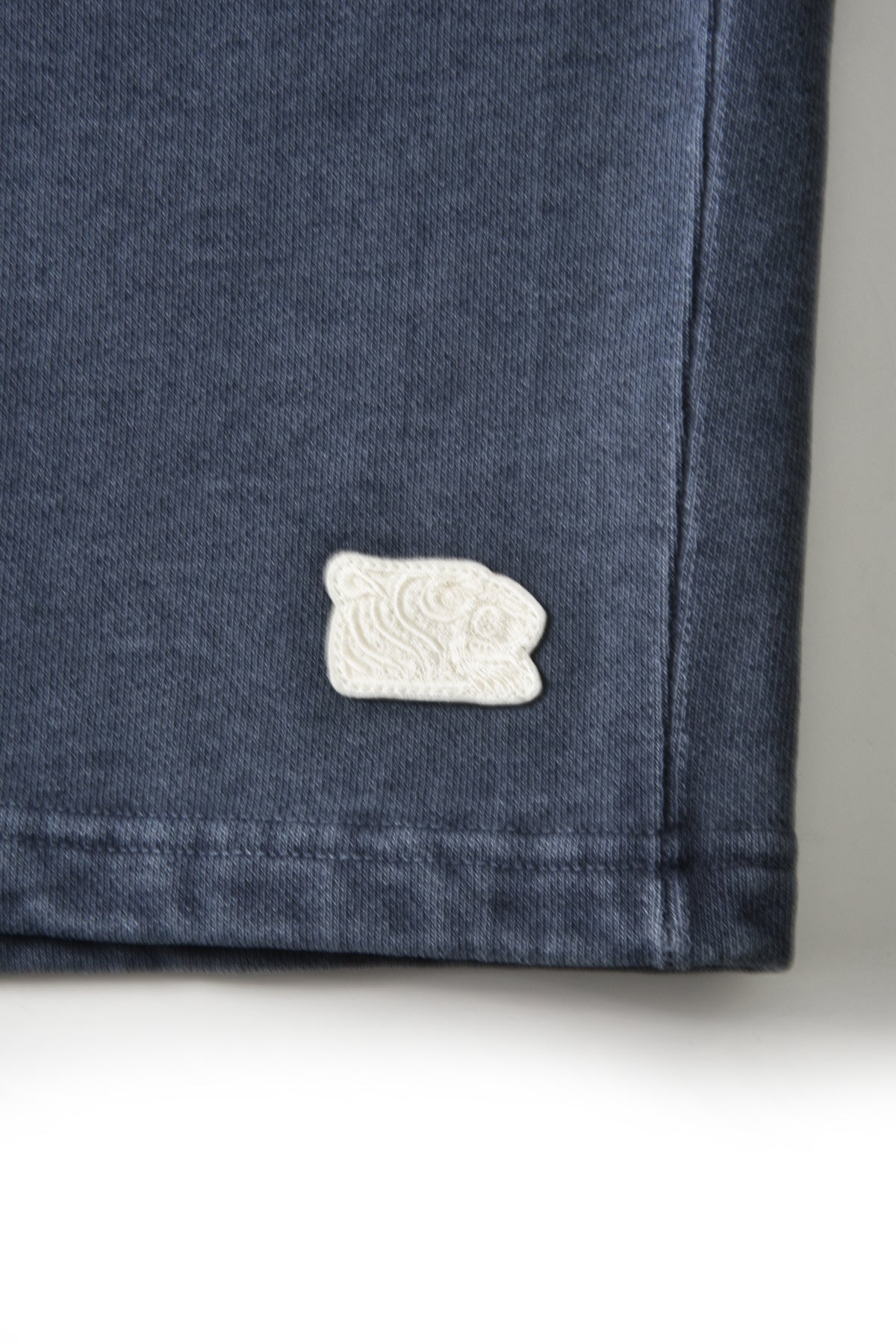 Close-up of a dark blue denim garment corner with a small white tiger head embroidered patch featuring a stylized face.